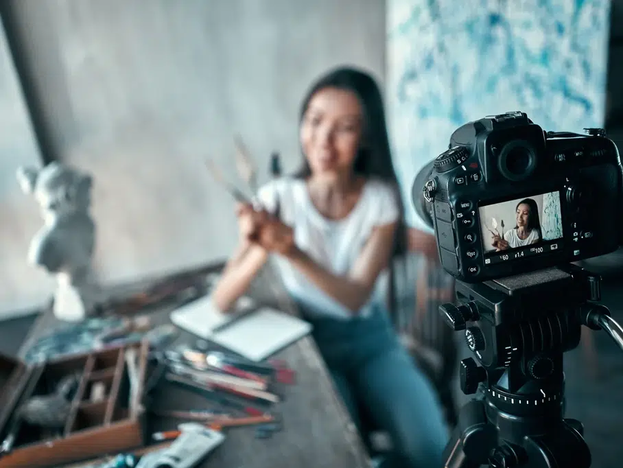 A person sits at a cluttered desk with art supplies, holding paintbrushes and discussing digital marketing strategies to the camera. The camera screen shows the person in focus, while a white sculpture is visible in the background.