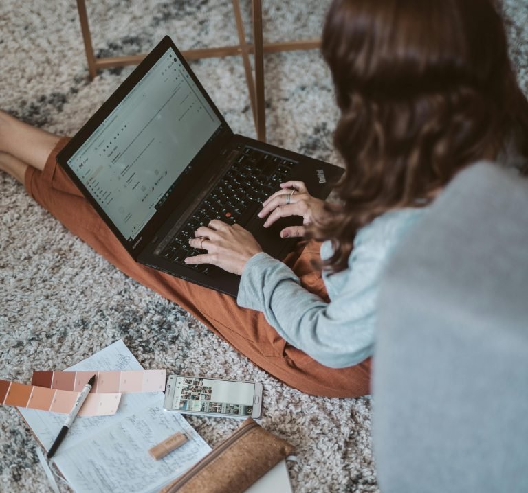 A person is sitting on a carpeted floor, working on a laptop. Nearby are color swatches, a phone, and scattered papers with notes on digital marketing strategies.
