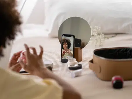 A person is applying makeup while looking at a smartphone, likely watching a digital marketing tutorial, placed on a wooden stand. Various makeup products and a cosmetic bag are spread out on a bed.