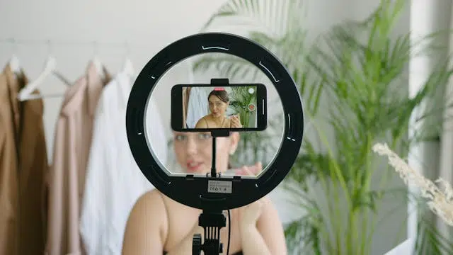 A person is recording a digital marketing video using a smartphone mounted on a ring light, with clothes hanging in the background and plants nearby.