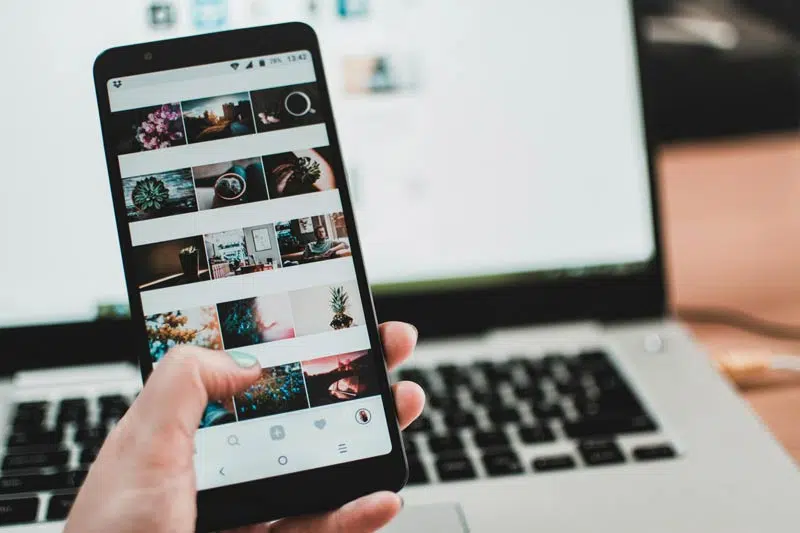 social media strategy - A person holds a smartphone displaying a social media app with photo thumbnails, showcasing the impact of digital marketing. The phone is held over an open laptop.