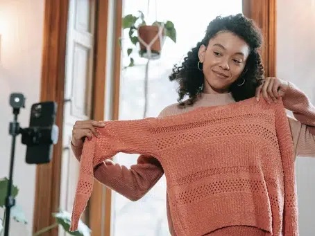 A person holds up a pink knitted sweater in front of a smartphone on a tripod, presumably filming for digital marketing purposes. The background showcases a window with a hanging plant, adding a touch of nature to the scene.