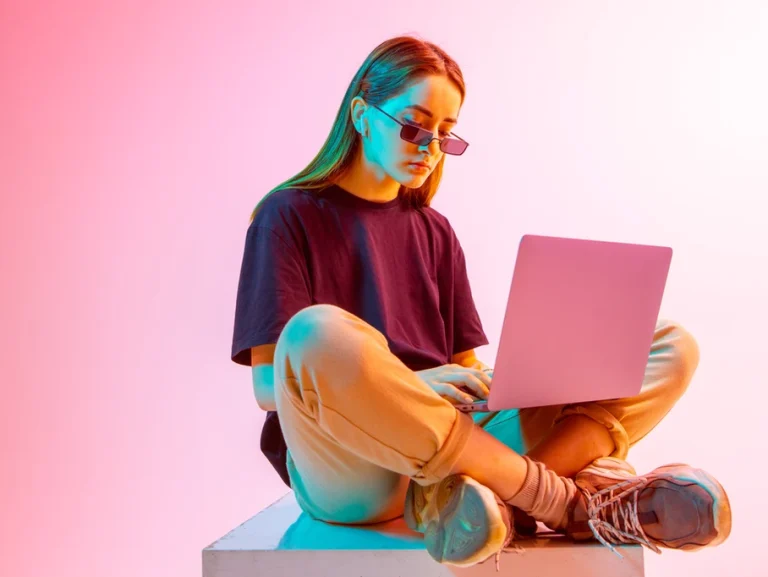 A person wearing sunglasses and casual clothing sits cross-legged on a platform, using a laptop for digital marketing, with a gradient pink and purple background.