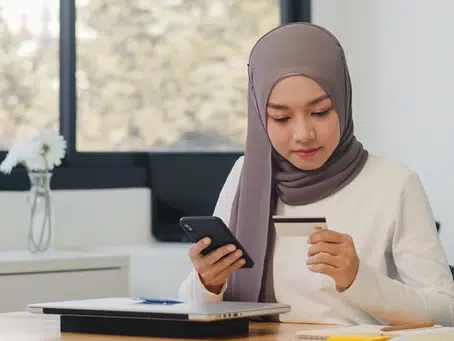 A woman in a hijab sits at a desk holding a smartphone and a credit card, with a laptop in front of her, engaging in digital marketing.